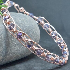 May include: A wire-wrapped bracelet crafted from rose gold-toned metal wire, featuring a series of faceted, translucent purple crystal beads secured within the intricate, twisted metal framework that forms a continuous, circular band design.