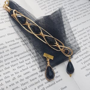 May include: A gold-toned wire-wrapped hair accessory featuring three faceted black teardrop beads encased in a lattice design, accompanied by a matching gold-toned hair pin with a single dangling black teardrop bead and black tulle fabric.