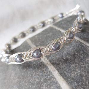 May include: A silver-toned wire-wrapped bracelet featuring a series of round, dark grey pearls. The metallic wire is intricately coiled around each individual pearl, creating a continuous, repeating pattern that forms the structure of the bracelet.
