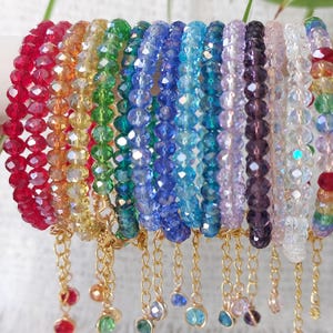 May include: A collection of multi-coloured beaded bracelets featuring faceted glass beads in a spectrum of hues including red, orange, yellow, green, blue, purple, and clear. Each bracelet includes a gold-toned metal chain extender with a bead charm.