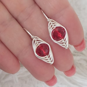 May include: Handmade earrings with a bright red gemstone set within a silver wire frame. The earrings have a teardrop shape and a hook fastening. These earrings are a striking accessory, adding a touch of elegance and colour.