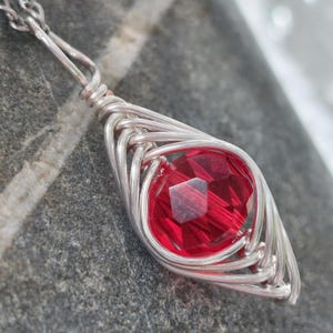 May include: A silver pendant necklace with a faceted, deep red gemstone. The gemstone is set within a woven silver wire setting, hanging from a delicate silver chain. The pendant is teardrop-shaped, with the red stone as the central feature.
