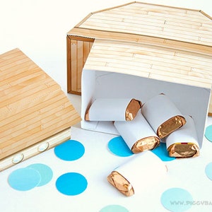May include: A wooden ark-shaped box with a hinged lid, partially open, revealing several wrapped candies. The box is made of light brown wood-grain patterned paper. Blue confetti dots the white surface. The website address "www.piggybankparties.com" is visible.