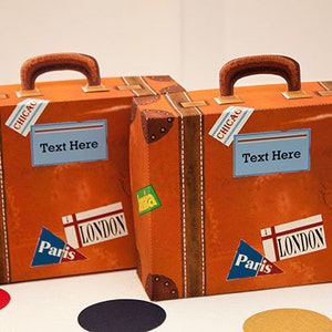 Puede incluir: Dos maletas decorativas naranjas con asas y protectores de esquina marrones. Cada maleta tiene pegatinas de viaje con los textos "Chicago", "London" y "Paris". El texto "Text Here" se muestra en una etiqueta azul en cada maleta.