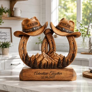 Könnte beinhalten: Eine dekorative Holzskulptur mit zwei Cowboyhüten auf zwei ineinandergreifenden Hufeisen. Die Hüte sind mit den Namen MASON und CHLOE graviert. Der Sockel zeigt ein Berg- und Waldrelief mit dem Text Adventure Together 21.06.2027.