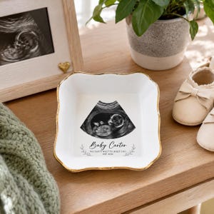 Könnte beinhalten: Eine quadratische weiße Keramikschale mit einem goldfarbenen, gewellten Rand. In der Mitte befindet sich ein schwarz-weißes Ultraschallbild über dem Text Baby Carter, We can't wait to meet you, und Est. 2026, eingerahmt von zwei kleinen Blättern.