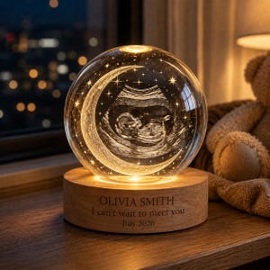 May include: A clear glass sphere featuring an internal 3D laser-etched ultrasound image of a fetus framed by a crescent moon and stars. The sphere rests on a circular wooden base engraved with the text OLIVIA SMITH, I can't wait to meet you, July 2026.