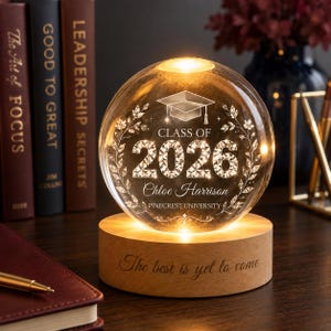 May include: A clear glass sphere engraved with a graduation cap, the text "CLASS OF 2026", "Chloe Harrison", and "PINECREST UNIVERSITY" surrounded by leaf motifs. It sits on a round wooden base engraved with the text "The best is yet to come".