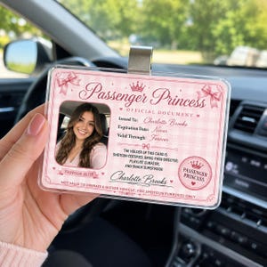 Könnte beinhalten: Ein rechteckiger Ausweishalter aus Kunststoff mit einer rosa-weiß karierten Scherzkarte mit der Aufschrift Passenger Princess. Die Karte enthält ein Foto, den Namen Charlotte Brooks sowie den Text Shotgun Certified und Forever.