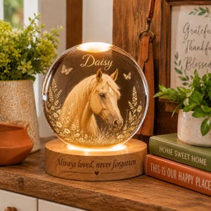 May include: A spherical glass ornament featuring an etched horse portrait, the name Daisy, and butterflies, illuminated from a circular wooden base. The base is engraved with the text Always loved, never forgotten and a small heart symbol.