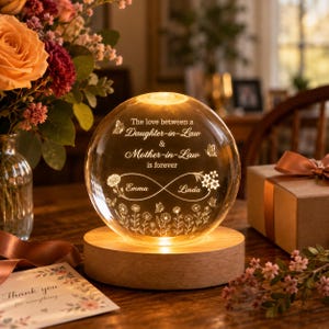 May include: A spherical glass night light on a circular wooden base, illuminated from within. The glass features an engraved infinity symbol, floral patterns, butterflies, and the text: The love between a Daughter-in-Law & Mother-in-Law is forever.