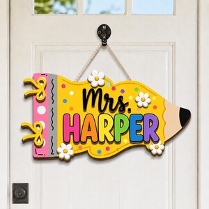 Könnte beinhalten: Ein gelbes, bleistiftförmiges Türschild aus Holz mit einem rosa Spiralbindungsabschnitt und gelben Akzenten. Der Text Mrs. HARPER ist in schwarzen und bunten Buchstaben aufgedruckt. Das Schild ist mit weißen Gänseblümchen und bunten Punkten verziert.