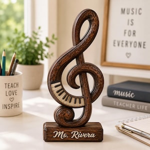 Könnte beinhalten: Eine dunkelbraune Holzskulptur in Form eines Violinschlüssels mit einer Klaviertastatur-Einlage entlang der Kurve. Die Skulptur steht auf einem rechteckigen Sockel mit dem eingravierten weißen Text "Ms. Rivera" auf der Vorderseite.