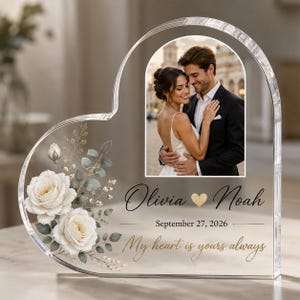 Könnte beinhalten: Eine herzförmige, transparente Acrylplatte mit einem aufgedruckten Foto eines Paares, weißen Rosen mit grünen Blättern sowie dem Text Olivia, Noah, September 27, 2026 und My heart is yours always in schwarzer und goldener Schreibschrift.