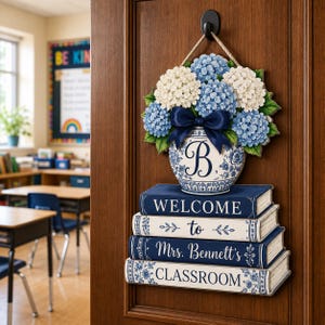Könnte beinhalten: Ein hängendes Türschild mit einer blau-weißen Vase voller blauer und weißer Hortensien, gekrönt von einer dunkelblauen Schleife. Unter der Vase befinden sich vier gestapelte Bücher mit dem Text: Welcome to Mrs. Bennett's CLASSROOM.