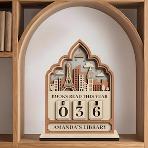 May include: A wooden bookshelf decor piece with a layered design featuring city skylines, the Eiffel Tower, and Big Ben. It displays "BOOKS READ THIS YEAR" with the number "036" and "AMANDA'S LIBRARY" at the bottom.