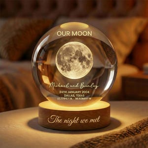 May include: A clear glass sphere with a detailed moon image inside, resting on a wooden base. The sphere is engraved with "OUR MOON", names, date, and location. The base reads "The night we met".