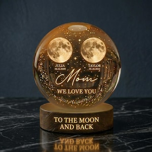 May include: A glowing glass sphere with two moon images, personalized with names and dates. The text "Mom, We Love You" is displayed, along with "To The Moon And Back" on the wooden base. The sphere is illuminated with a warm light.