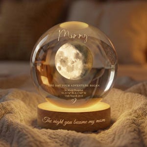 May include: A clear glass sphere with a moon design, resting on a wooden base. The sphere has the word "Mummy" and the text "The day your adventure began" engraved on it. The base has the text "The night you become my mom".