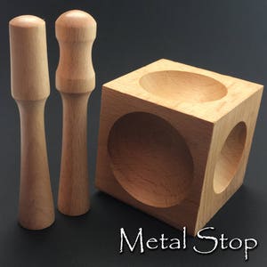 Könnte beinhalten: Zwei Holzhammer mit abgerundeten Enden und ein Holzwürfel mit drei runden Vertiefungen. Der Würfel ist mit "Metal Stop" beschriftet.