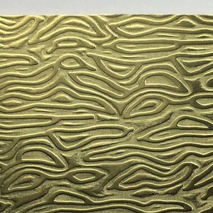 May include: A rectangular brass-colored metal plate with a textured, flowing pattern. The surface features a series of curved, parallel lines, creating a wave-like appearance. The text "Metal Stop" is printed in black at the bottom.