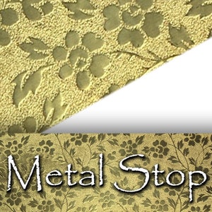 Op de afbeelding: Close-up van een goudkleurig metalen oppervlak met een bloemenpatroon. Het ontwerp heeft in reliëf aangebrachte bloemen en bladeren. De tekst "Metal Stop" is onderaan zichtbaar. Het algemene thema is decoratief metaalwerk.