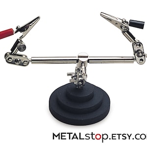 May include: A silver-colored helping hands tool with two adjustable alligator clips, one with a red cap and the other with a black cap. The tool is mounted on a black, tiered base. The text "METALstop.ETSY.com" is visible.