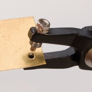 Puede incluir: Una herramienta de mano de metal negro con un punz&oacute;n de metal plateado utilizado para crear un agujero en una pieza de lat&oacute;n.