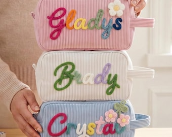 May include: Three rectangular corduroy pencil cases stacked vertically. The top case is pink with the name Gladys in colorful yarn. The middle case is white with the name Brady in green yarn. The bottom case is light blue with the name Crystal in colorful yarn and small flower appliqués.