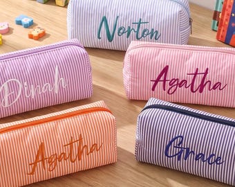 Op de afbeelding: Vijf rechthoekige etuis met gestreept patroon in lichtblauw, paars, roze en oranje. Elk etui is voorzien van een gepersonaliseerde naam geborduurd in cursief schrift: Norton, Dinah, Agatha en Grace. De etuis hebben een ritssluiting.