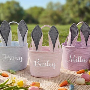 May include: Three Easter baskets with bunny ears. Each basket is a different pastel color: blue, pink, and purple. The baskets have white handles and are personalized with names. The bunny ears are black and white checkered fabric.