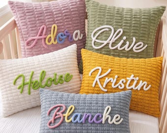 Op de afbeelding: Vijf rechthoekige en vierkante kussens met een textuur in mauve, olijfgroen, crème, mosterdgeel en leiblauw. Elk kussen is voorzien van een verhoogde, zacht aanvoelende naam in cursief schrift: Adora, Olive, Heloise, Kristin en Blanche.