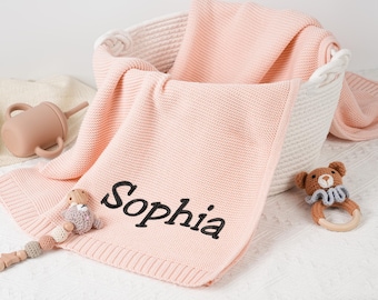 May include: A pink knitted baby blanket with the name "Sophia" embroidered in black. The blanket is folded in a white basket, with a matching sippy cup and a wooden and crochet toy.