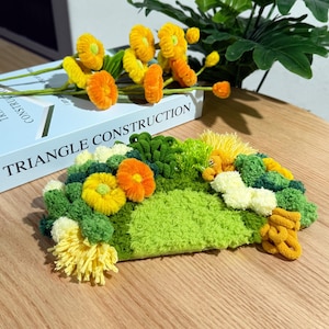 Könnte beinhalten: Ein farbenfrohes, strukturiertes Kunstwerk mit grünen, gelben und orangen Garnen, das einer Landschaft ähnelt. Das Stück zeigt verschiedene Texturen und Farben. Der Text "TRIANGLE CONSTRUCTION" ist auf einem Buch im Hintergrund zu sehen.