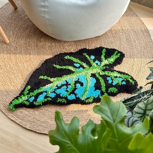 May include: A handmade, leaf-shaped rug with a black background and a detailed pattern in shades of green and blue. The rug is placed on a natural fiber round rug, with a white beanbag chair in the background. The rug's design resembles a tropical plant leaf.