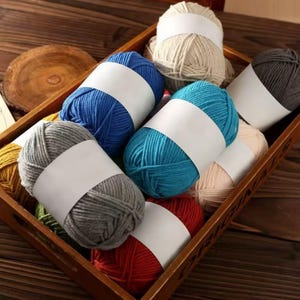 May include: A wooden tray filled with colorful yarn skeins. The yarn is in various colors, including blue, teal, gray, red, and beige, each wrapped with a white label. The tray sits on a wooden surface.