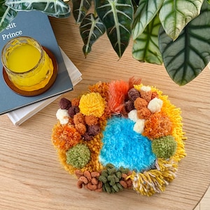 Könnte beinhalten: Ein farbenfrohes, strukturiertes Textilkunstwerk mit einem zentralen türkisfarbenen Bereich, der von verschiedenen Texturen und Farben umgeben ist, darunter Gelb, Orange und Braun. Das Werk liegt auf einer Holzoberfläche, mit einem Glas gelber Flüssigkeit und einem Buch im Hintergrund.