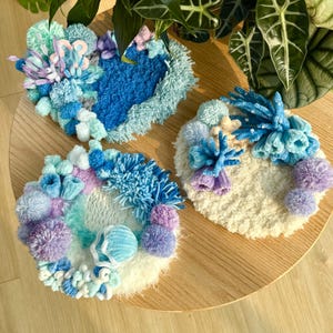 May include: Three handmade, textured fiber art pieces in the shape of a heart and circles. The pieces feature a variety of blue, purple, and white yarn, with pom-poms and other decorative elements. The pieces are displayed on a wooden surface.