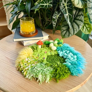 Può includere: Un'opera d'arte tessile fatta a mano che raffigura un paesaggio in miniatura. Presenta varie trame e sfumature di verde, giallo e blu, con un piccolo accento di fungo rosso. L'opera circolare è su un tavolo di legno.