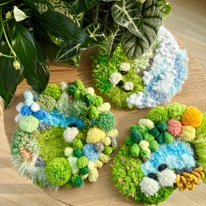 Op de afbeelding: Drie handgemaakte, ronde textielkunstwerken met een landschapsthema. Elk stuk heeft een verscheidenheid aan texturen en kleuren, waaronder groen, blauw, geel en wit, die lijken op miniatuurlandschappen. De stukken zijn gerangschikt op een houten oppervlak.