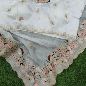 May include: White embroidered blouse with a matching sheer grey scarf. The blouse features floral embroidery in pink, brown, and gold. A small figurine sits on top of the scarf. The fabric has a delicate, flowing appearance.