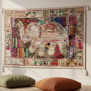 May include: A vibrant elephant tapestry hangs on a white wall. The elephant is cream-coloured with detailed patterns. The tapestry has a patchwork border with various colours and patterns. Two floor cushions and a side table with a plant and candle are in the foreground.