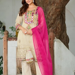 Könnte beinhalten: Beiges besticktes Tunika- und Hosen-Set mit Spiegelverzierungen. Die Tunika hat einen runden Ausschnitt und Dreiviertelärmel. Ein leuchtend rosa Schal liegt über der Schulter. Das Outfit wird mit passenden beigen Hosen und transparenten Sandalen kombiniert.