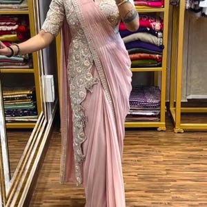 Puede incluir: Un sari rosa claro con intrincados bordados plateados en el borde y una blusa a juego. El sari está elegantemente drapeado, mostrando los intrincados adornos. La blusa presenta un cuello alto y mangas cortas con detalles plateados similares. El fondo incluye estantes de telas coloridas.