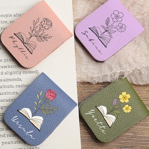 Może przedstawiać: Cztery skórzane zakładki w kolorze różowym, fioletowym, niebieskim i zielonym, każda z kwiatowym wzorem i imieniem. Zakładki przedstawiają ilustrację otwartej książki i leżą na drewnianej powierzchni.