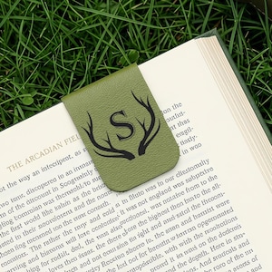 May include: An olive green magnetic bookmark featuring a black printed design of a capital letter S centered between two stylized antlers. The bookmark is clipped onto the top edge of an open book page with printed text visible on the paper.