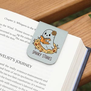 Op de afbeelding: Een lichtblauwe magnetische boekenlegger met een cartoon-geestje met een bloem op het hoofd, lezend in een oranje boek terwijl het in een nest van herfstbladeren zit. De tekst "SPOOKY STORIES" staat in het zwart onder de illustratie gedrukt.