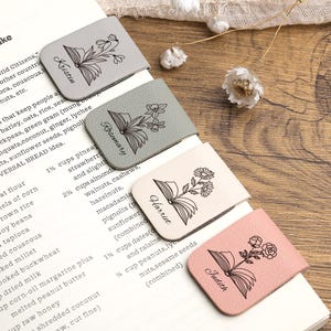 Puede incluir: Cuatro marcadores de cuero en gris, verde, crema y rosa, cada uno con un diseño floral y un nombre: Kristen, Rosemary, Harriet y Judith. Los marcadores están en una página de libro.