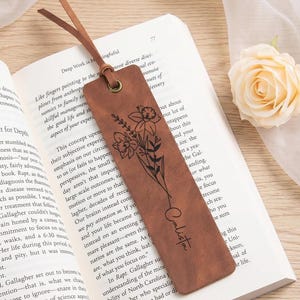 May include: A brown leather bookmark with a floral design and the name "Calista" printed on it. The bookmark has a leather loop and is placed in an open book.