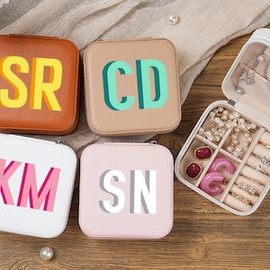 May include: An assortment of personalized jewelry boxes in various colors, including brown, beige, white, and pink. Each box features initials in bold, contrasting colors. One box is open, revealing jewelry.